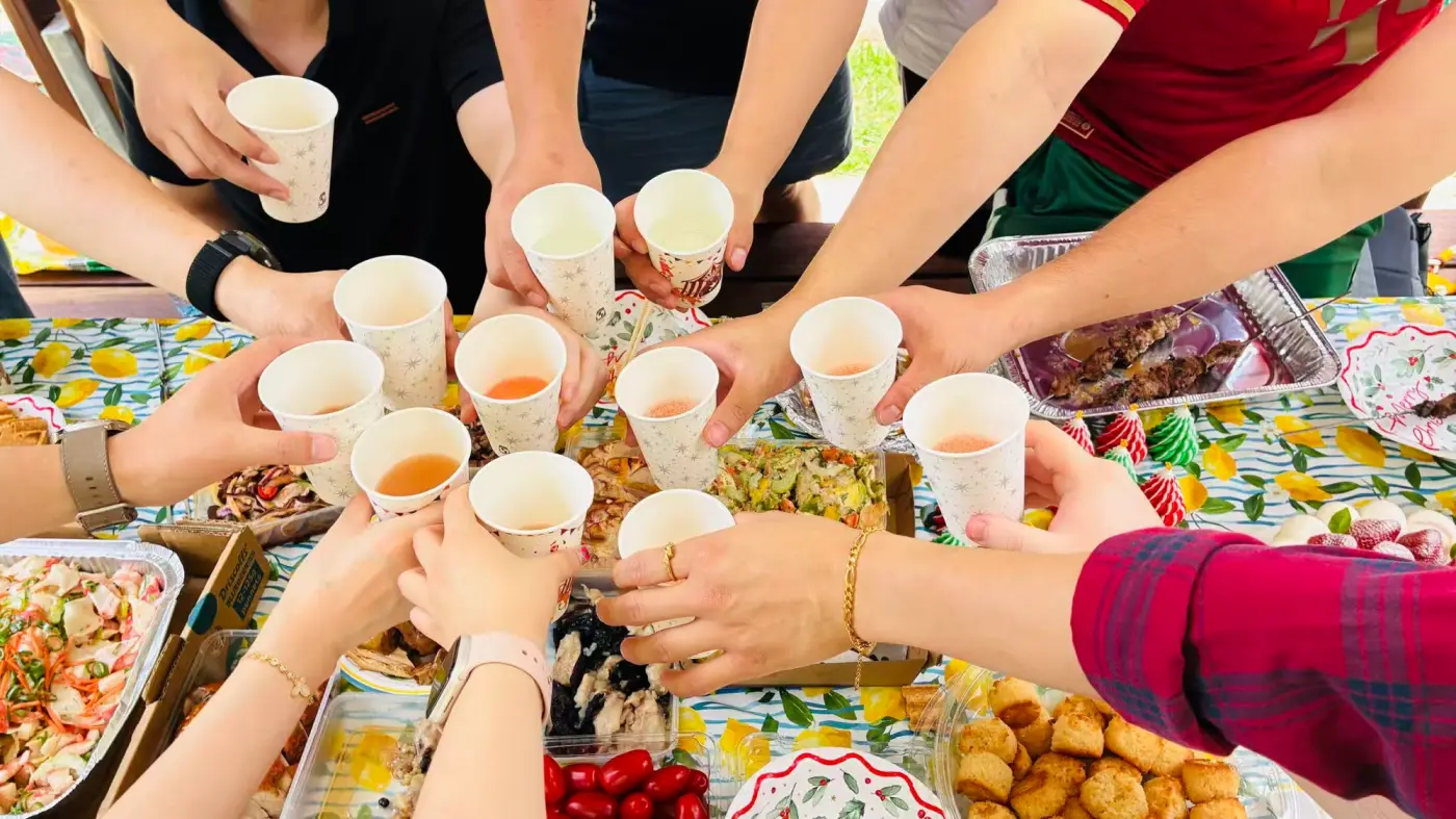 Everyone reaching in to clink cups over a shared table of food at the club picnic
